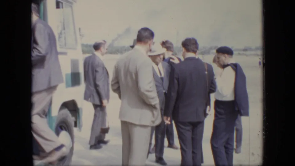 A group of men in suits and hats stand on sandy ground at the 1971 Moscow site, with a vehicle edge and industrial structures nearby.