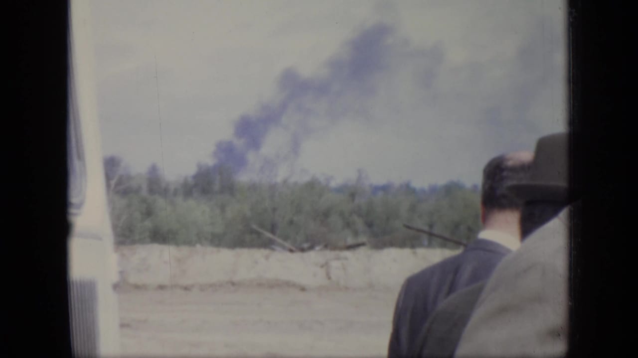 1971 Moscow home-movie frame with suited men partly turned away as a dark smoke plume rises over a treeline.