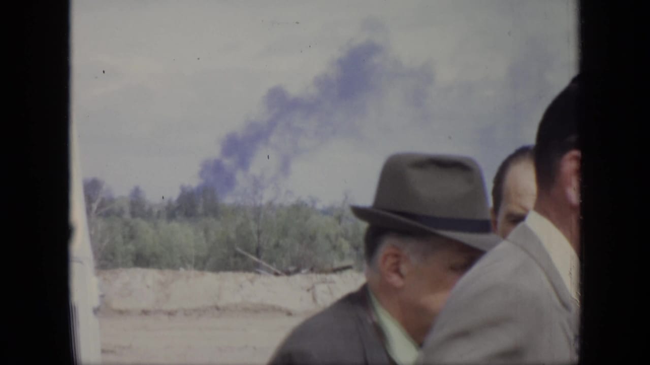 1971 Moscow frame showing adult men in suits and hats near a treeline, with a smoke plume visible in the background.