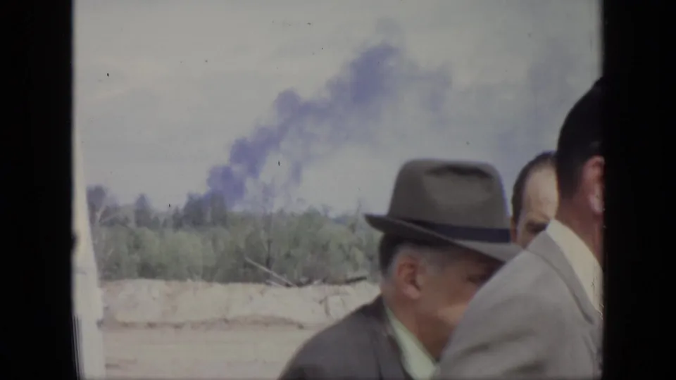 1971 Moscow frame showing adult men in suits and hats near a treeline, with a smoke plume visible in the background.