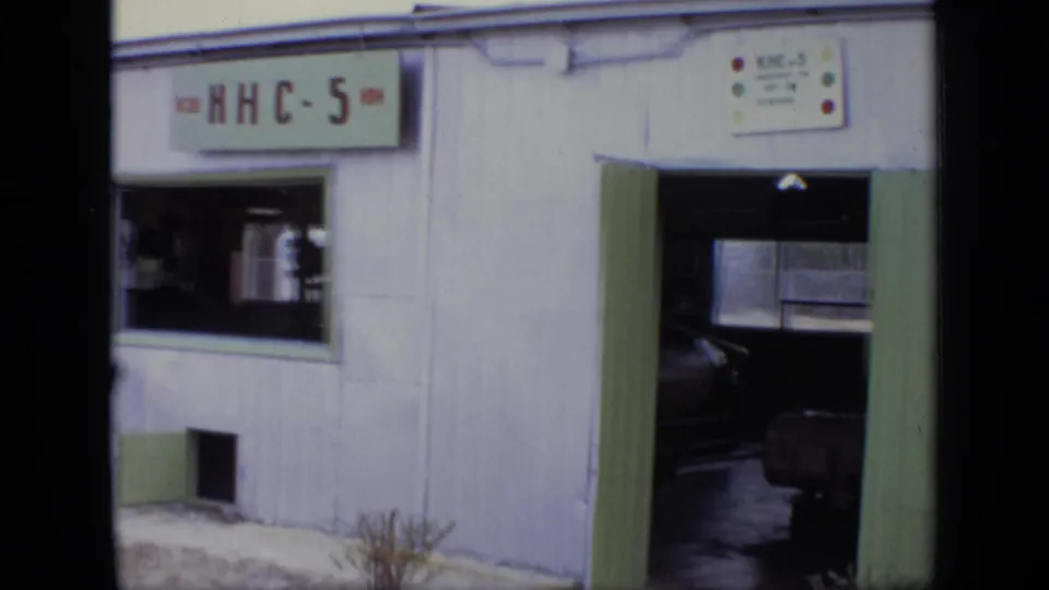 1971 Moscow frame of a small white building with green trim, an RHC-5 or KHC-5 sign, open doorway, window, and scrubby ground.