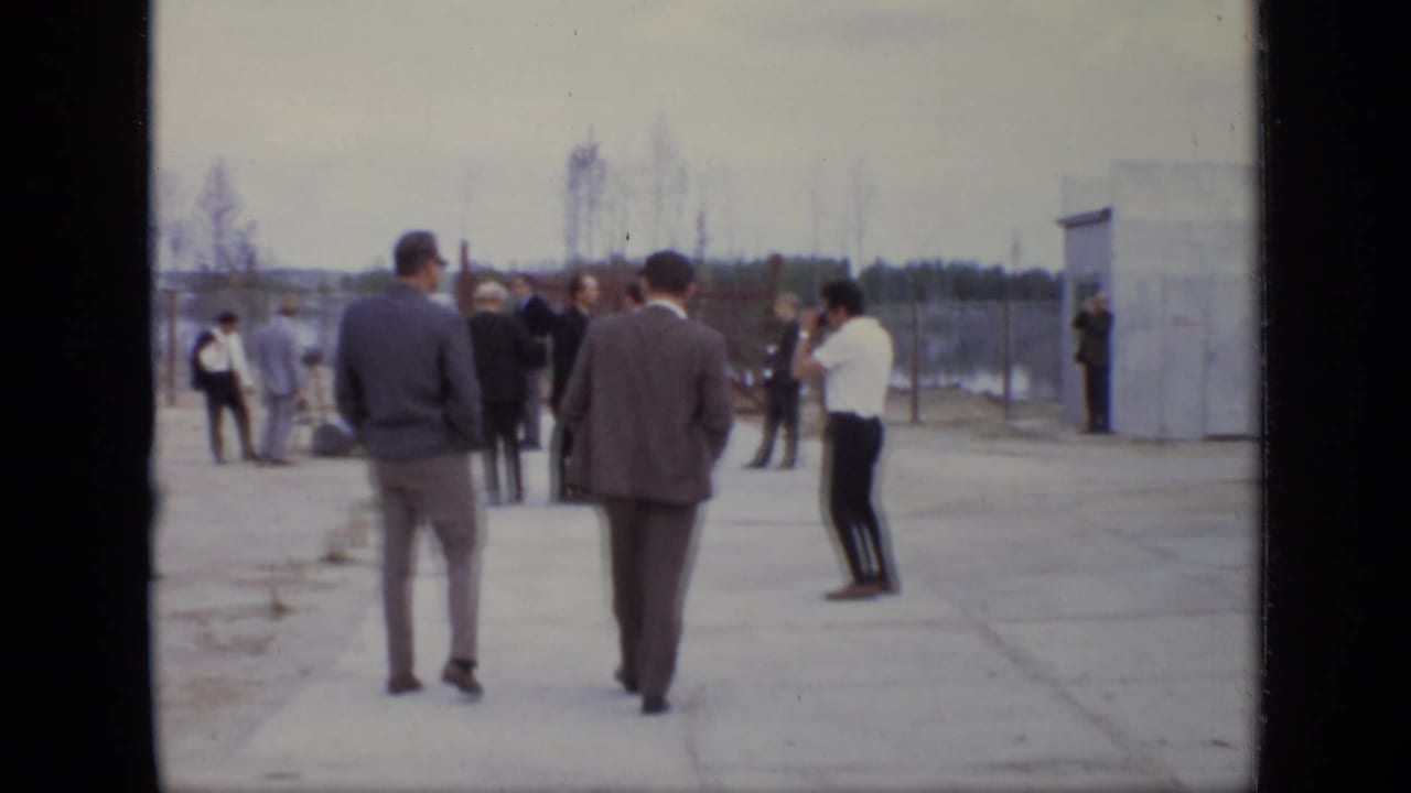 Men stand and walk in a paved industrial yard near a small prefabricated building.