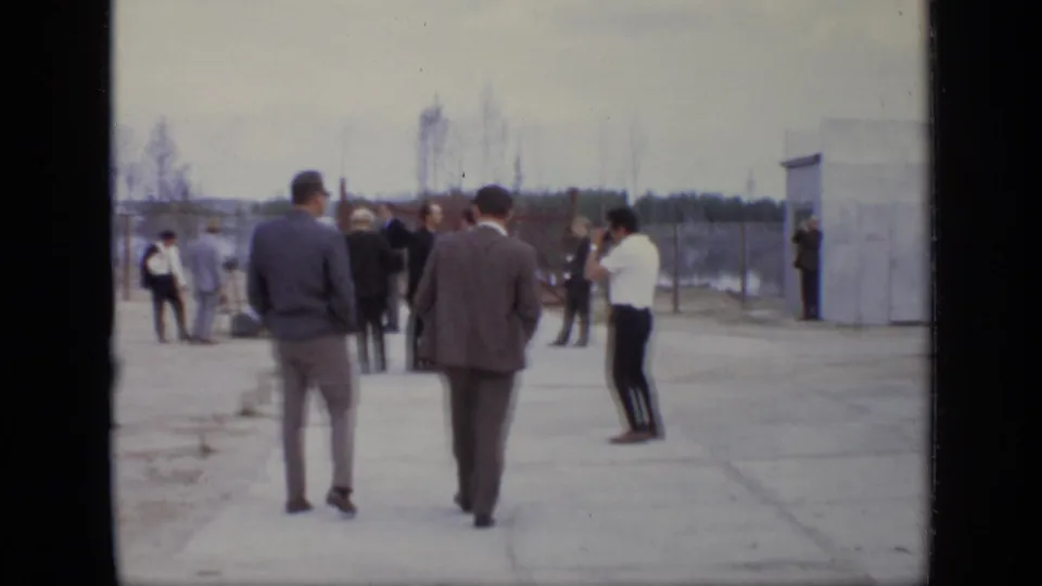 In Moscow, 1971, men in suits and casual clothing gather in an outdoor industrial yard near a small building.
