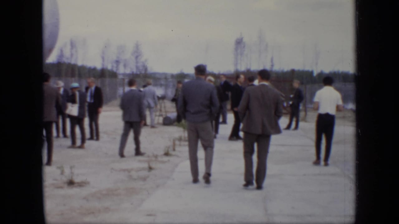 Group of men walk and stand across a paved industrial area under an overcast sky.