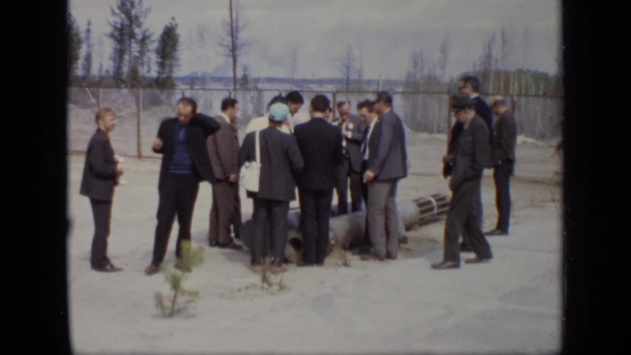 Group of formally dressed men at a 1971 Moscow industrial site, standing near a large pipe or equipment section by a fence.