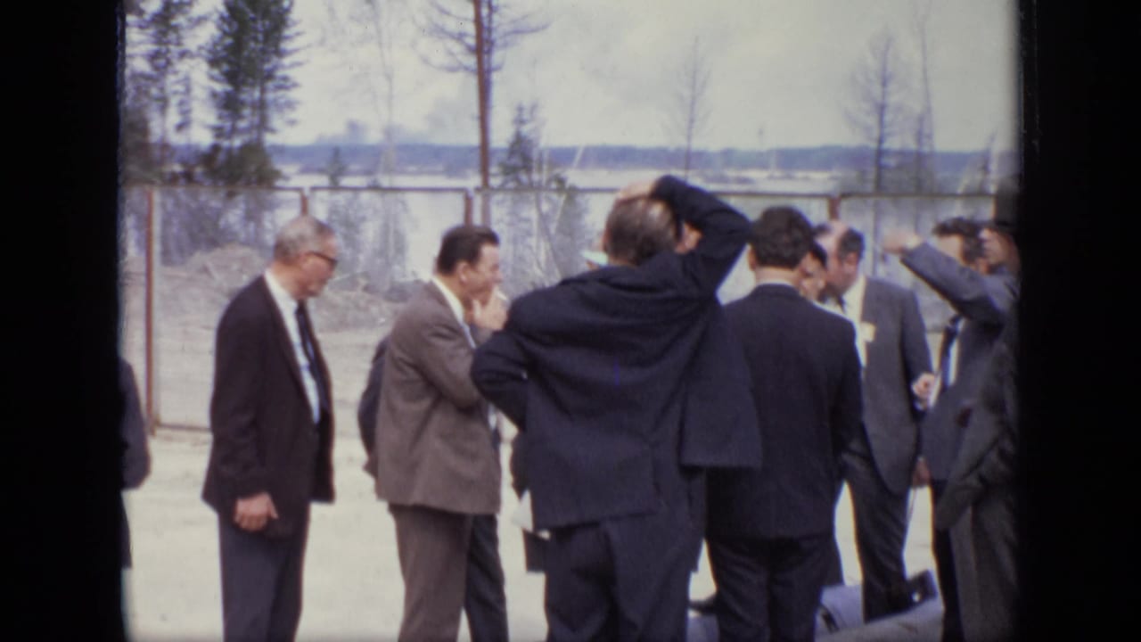 1971 Moscow home-movie frame showing adult men in suits gathered outdoors near a chain-link fence.