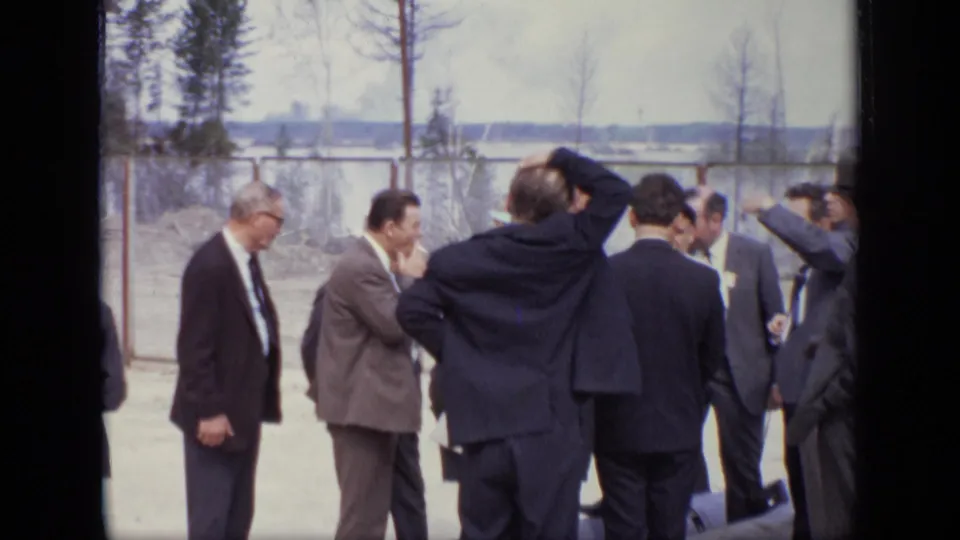 1971 Moscow home-movie frame showing adult men in suits gathered outdoors near a chain-link fence.