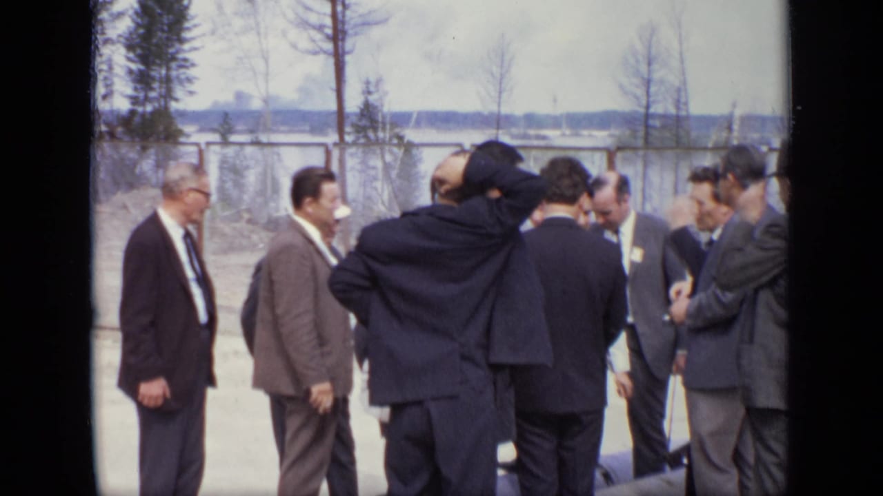 Group of suited men converse near a fence, with one man raising an arm near his head.