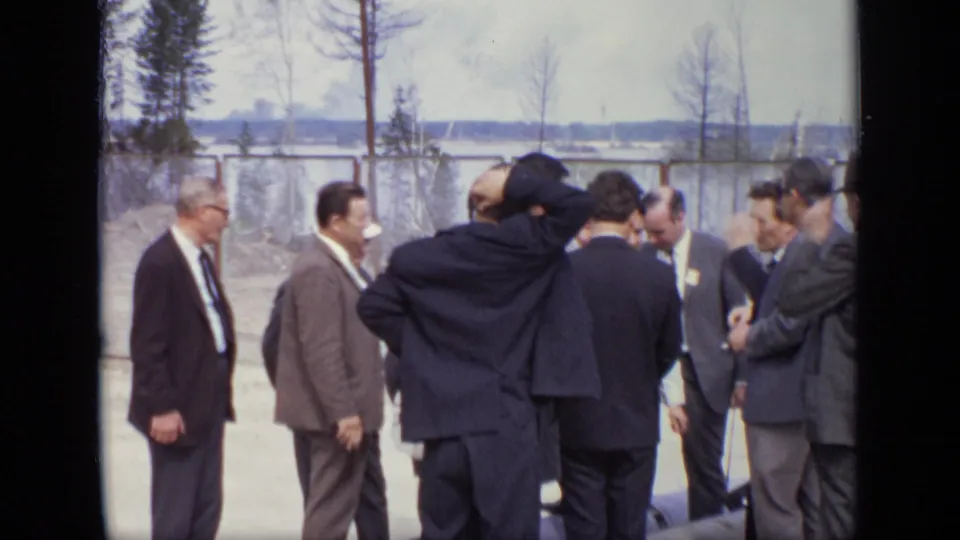 Adult men in 1971 Moscow stand in a loose cluster outdoors, wearing suits and ties near a fence.