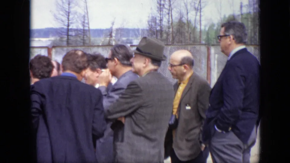 Closer 1971 Moscow frame of suited adult men, including a man in a fedora, gathered near a chain-link fence.