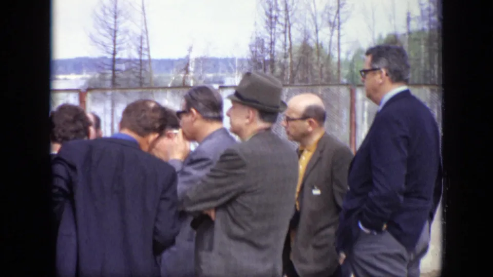 1971 Moscow home-movie frame with suited men seen from the side and back near a fenced outdoor area.