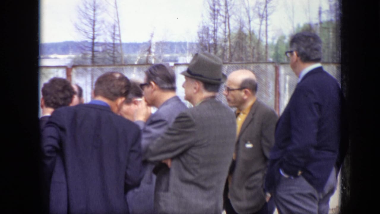 Adult men in suits, hats, and glasses gather outdoors in Moscow, 1971, near fencing and a distant waterside background.