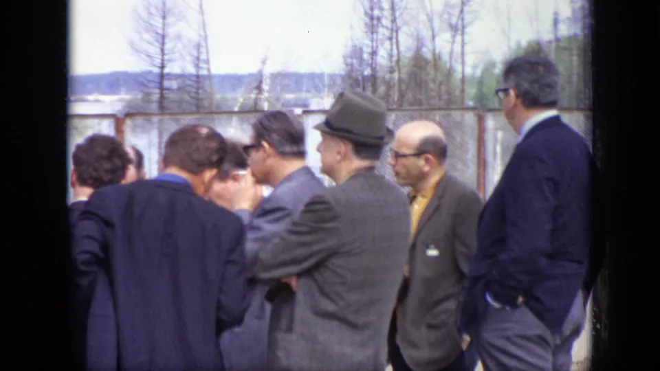 Adult men in suits, hats, and glasses gather outdoors in Moscow, 1971, near fencing and a distant waterside background.