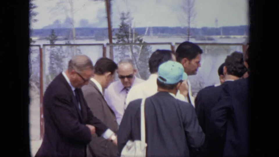 Adult men in 1971 Moscow stand in conversation near a chain-link fence, with sunglasses, suits, and a light cap visible.