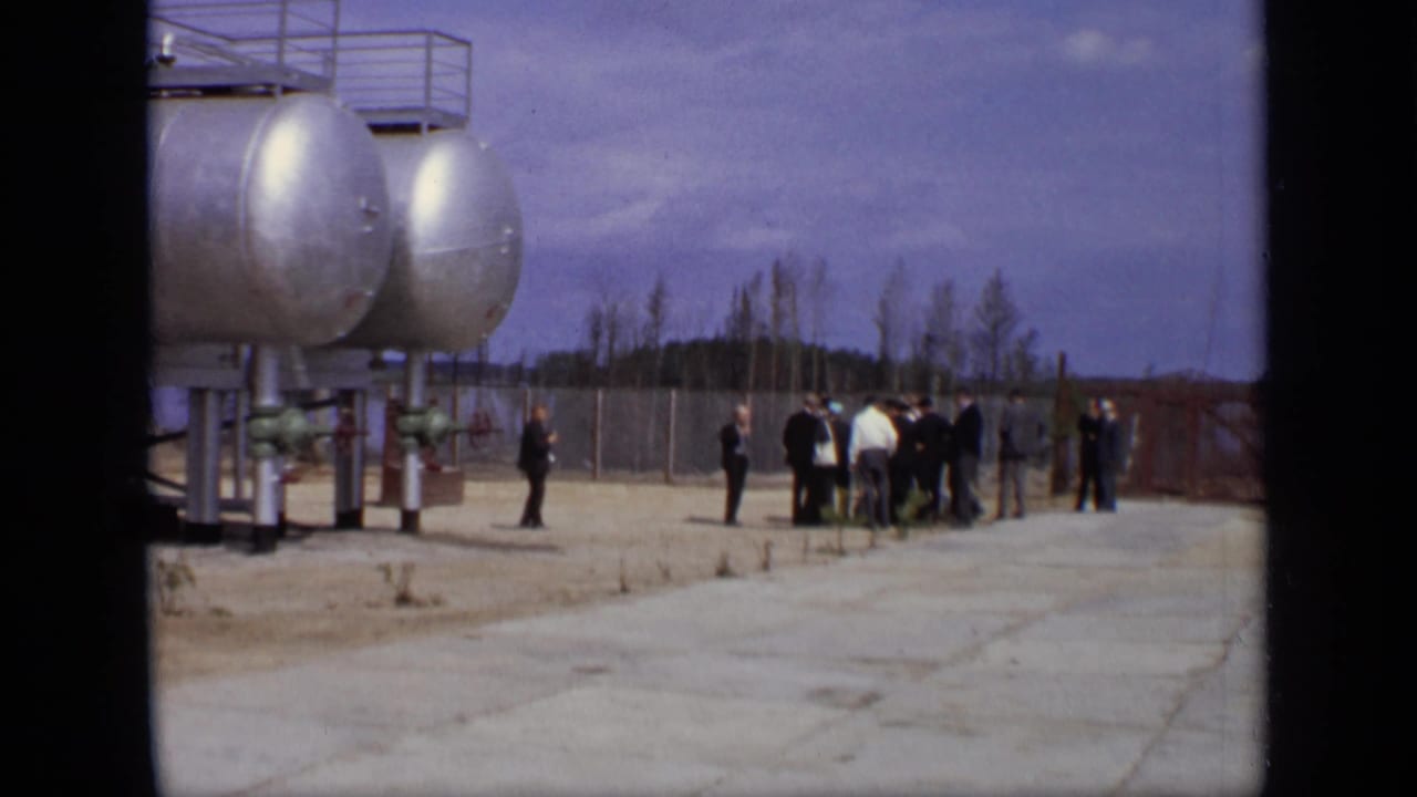 Men standing and walking near large silver storage tanks in a concrete industrial yard.