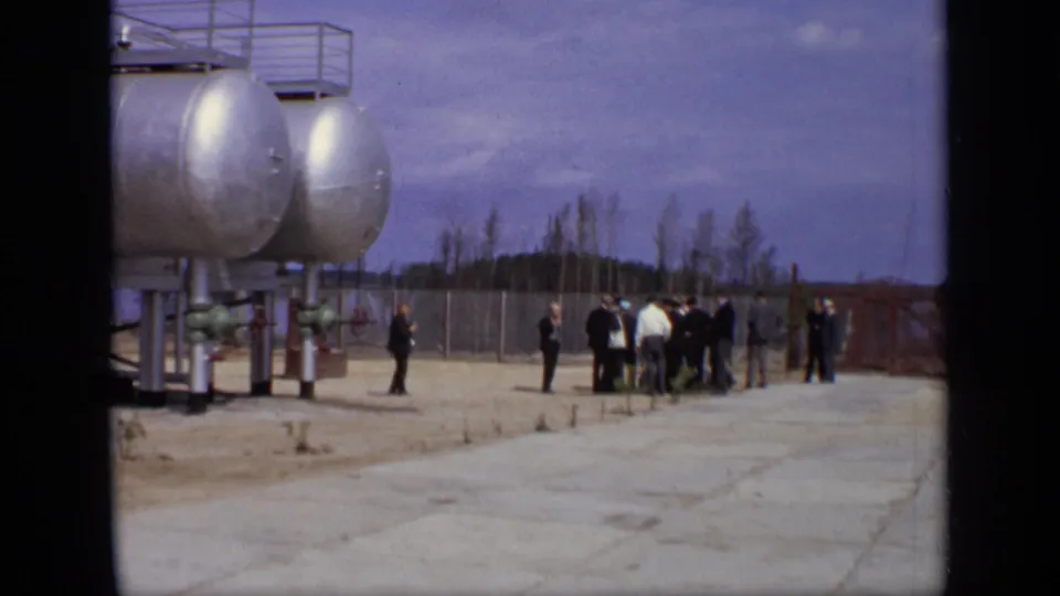 1971 Moscow home-movie frame showing suited men near large silver storage tanks inside a fenced industrial yard.