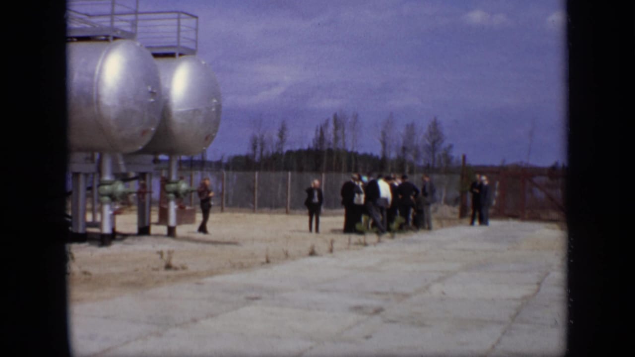 Group of men standing near tanker tanks and fencing in an open industrial yard.