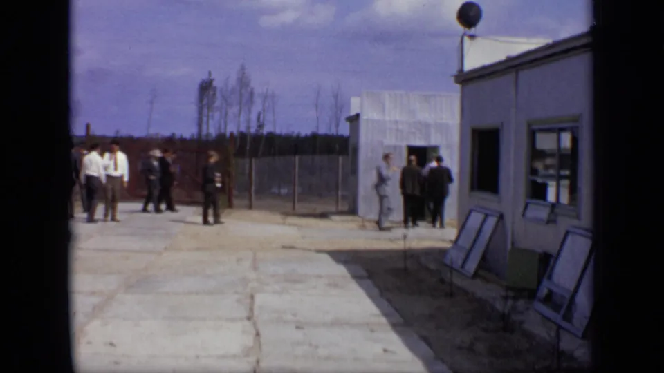 1971 Moscow frame with men in suits near a small white building and fenced industrial grounds.