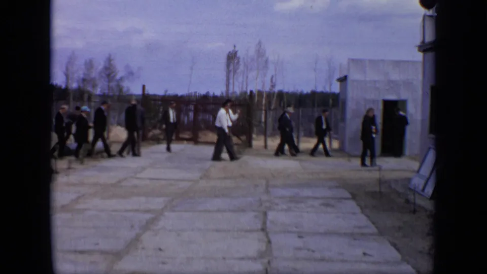 Men in suits walk across a Moscow industrial yard near a small building and perimeter fence in 1971 home-movie footage.