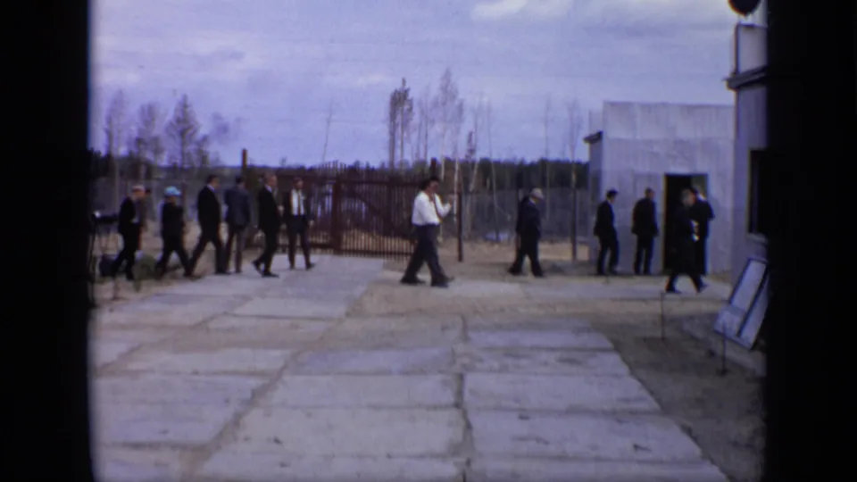 1971 Moscow 8mm frame showing men in dark suits crossing a concrete courtyard beside a small building and tall fence.