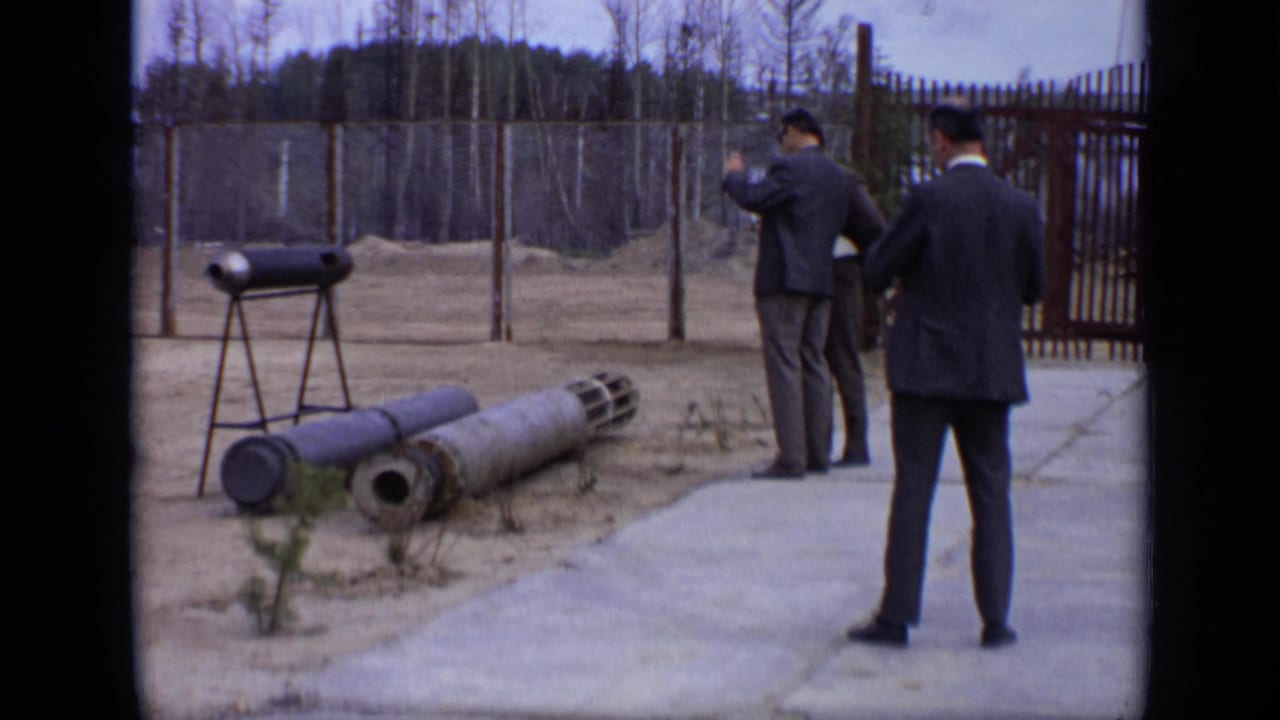 Two men stand near cylindrical objects displayed beside a fenced outdoor area.