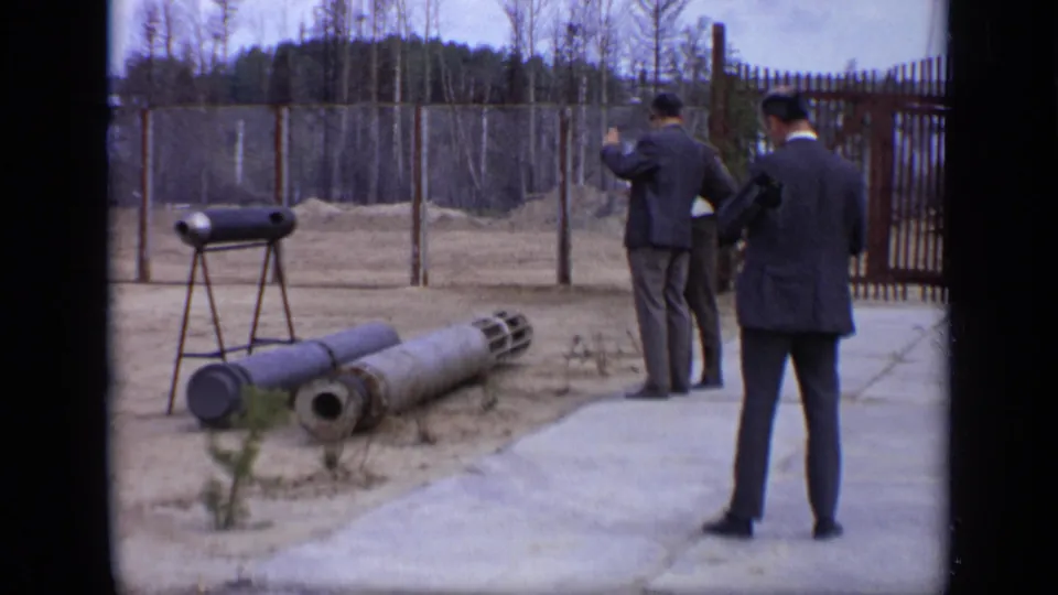 1971 Moscow 8mm frame showing men in suits near ordnance-like cylinders, a tripod-mounted object, fencing, and bare trees.