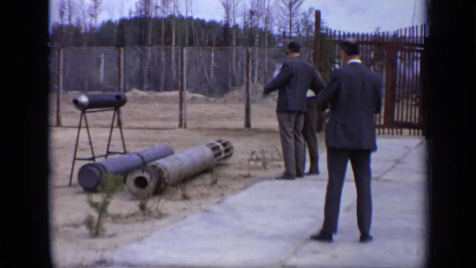 1971 Moscow 8mm frame with a suited man in the foreground and others near ordnance-like cylinders in a fenced courtyard.