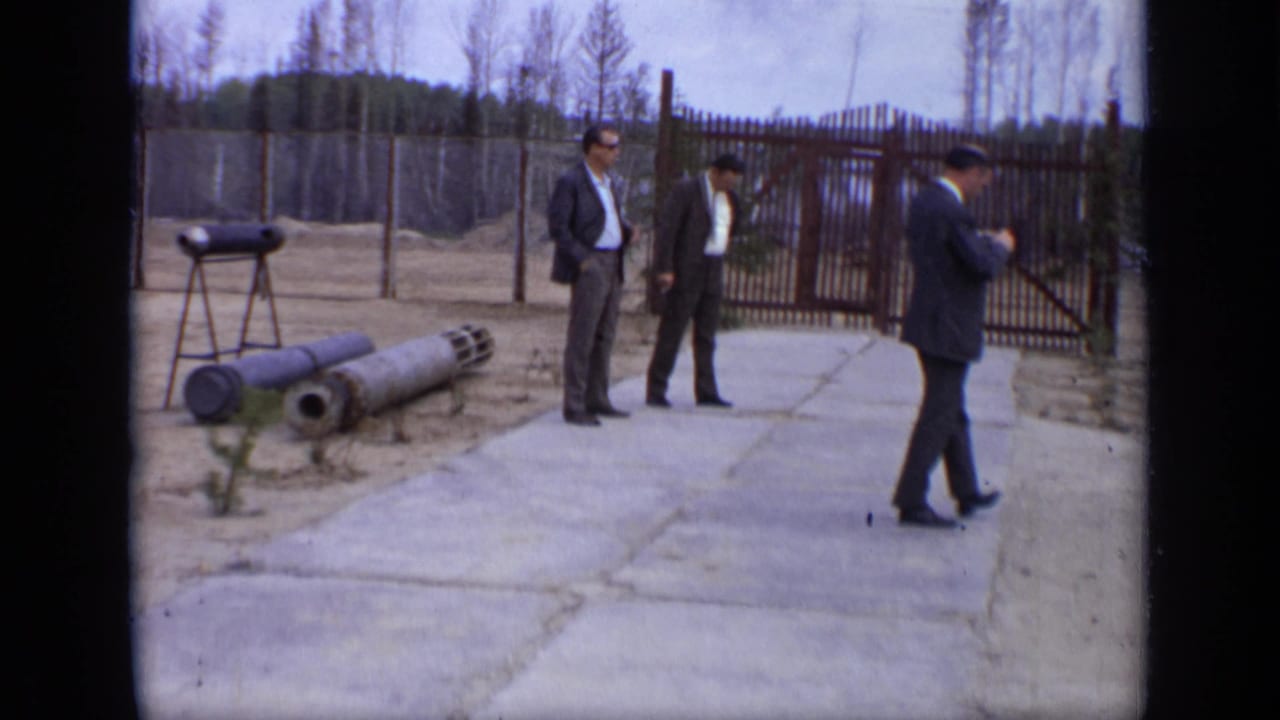 1971 Moscow 8mm frame showing three suited men near a fenced gate, concrete path, bare trees, and large cylindrical objects.