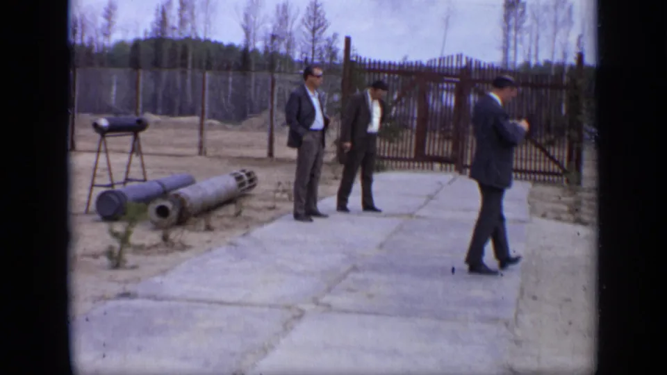 1971 Moscow 8mm frame showing three suited men near a fenced gate, concrete path, bare trees, and large cylindrical objects.