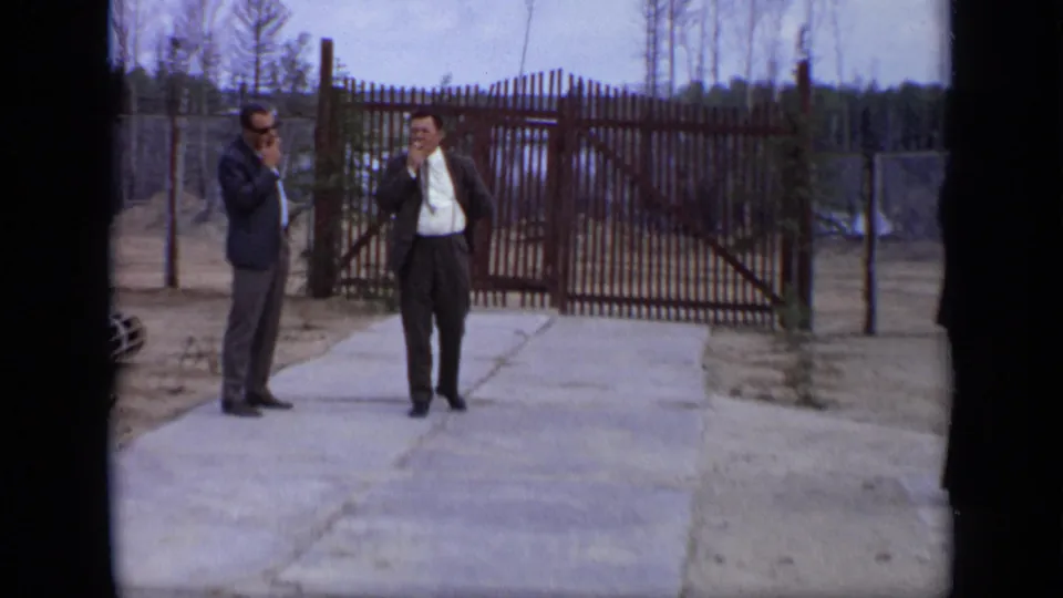 1971 Moscow 8mm frame showing two suited men in conversation or smoking beside a fenced compound gate.