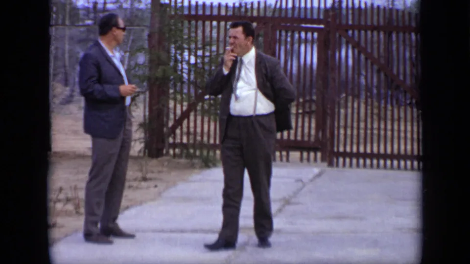1971 Moscow 8mm close frame of two suited men near a fenced gate, with one man appearing to smoke or hold a small object.