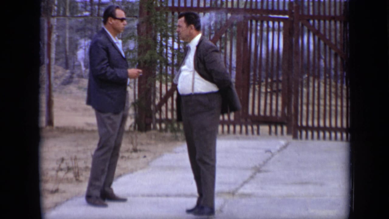 Two men in suits talk near a large wooden gate and a paved path.