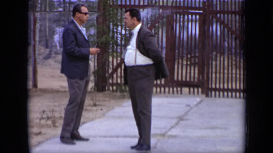Two adult men in suits, one wearing sunglasses, stand talking outdoors at a Moscow industrial site in 1971.