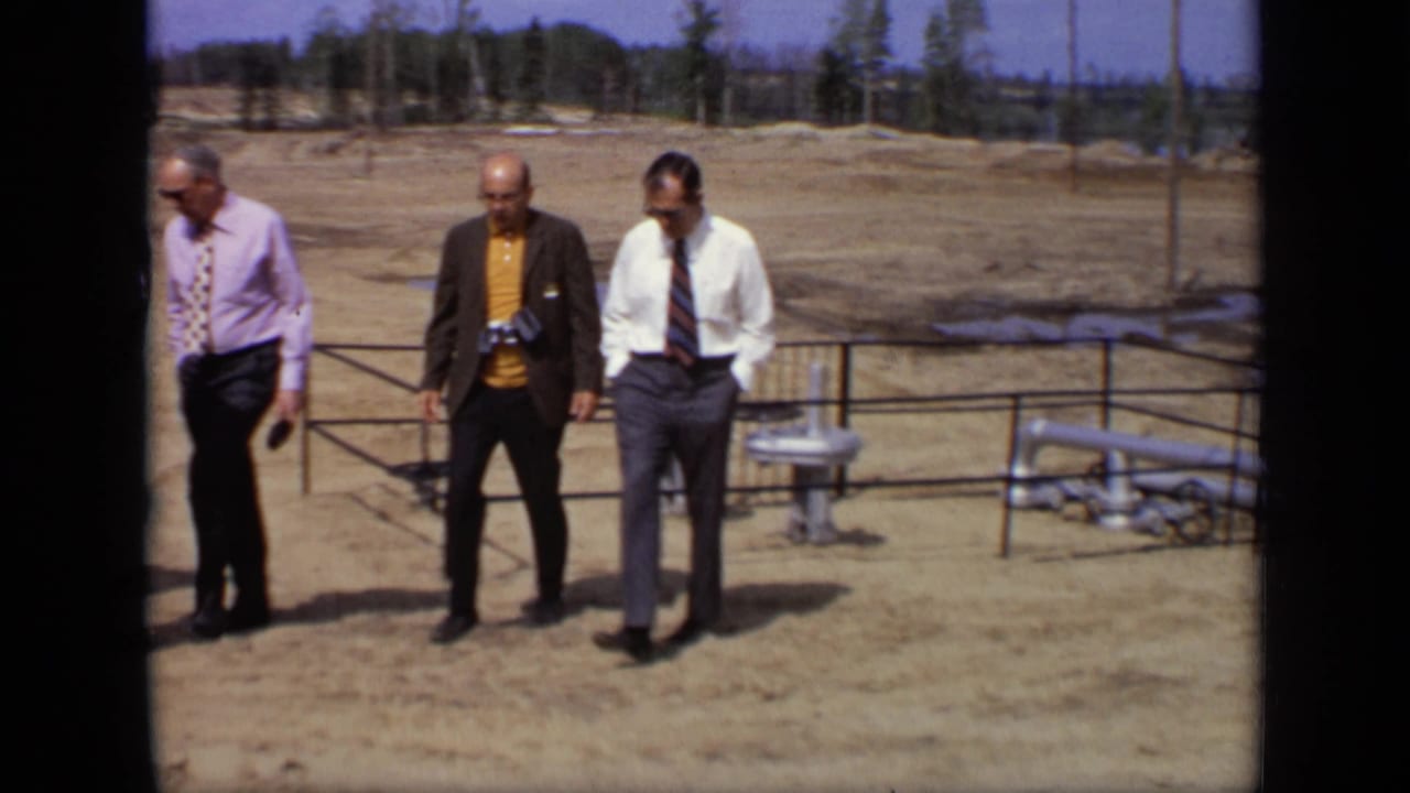 Three men in business attire walk near a railing and industrial piping in a dusty open field.