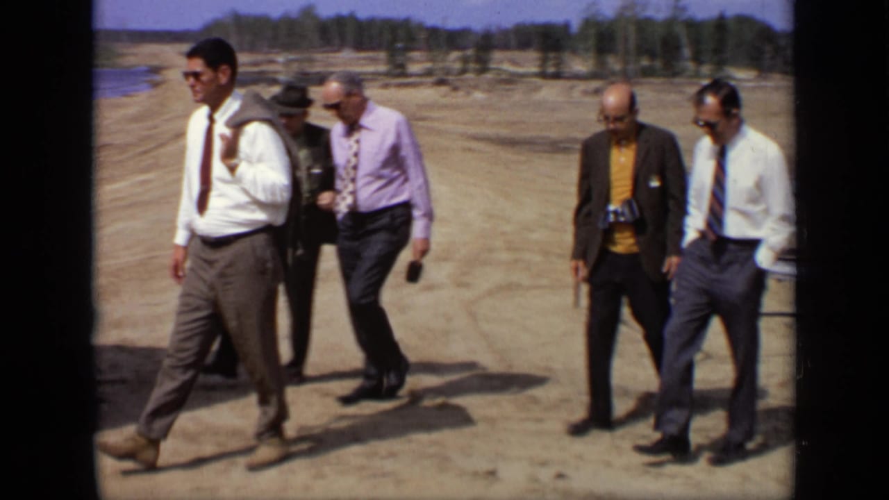 Five men in business attire walk across a dirt road beside water and a treeline.