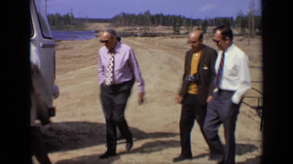 Three formally dressed men walk past the edge of a vehicle in a dusty Moscow field, with water and treeline visible in the background.
