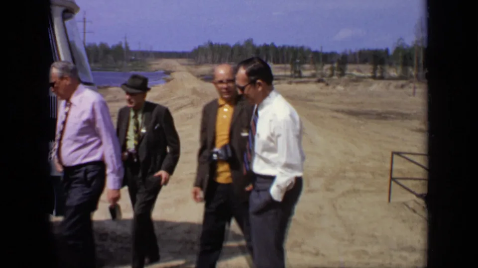 Four men in 1971 Moscow footage walk beside a parked vehicle, with a dusty open site, railing, water, and treeline in the background.