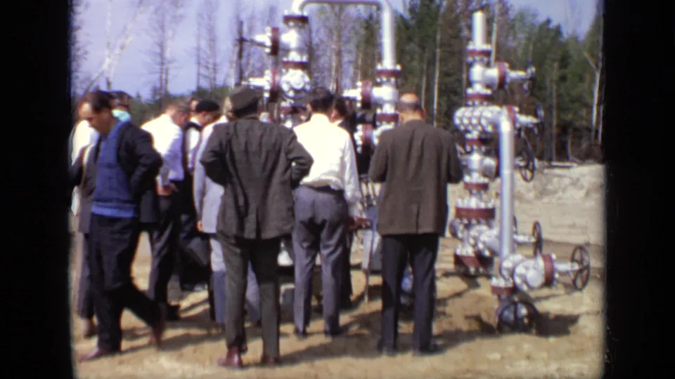 The 8mm frame shows men viewed mostly from behind as they examine outdoor piping and valve hardware in a wooded work area.