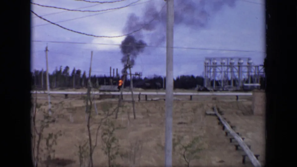 1971 Moscow-area 8mm frame with a small burning structure, black smoke, dry ground, utility poles, and distant industrial framing.