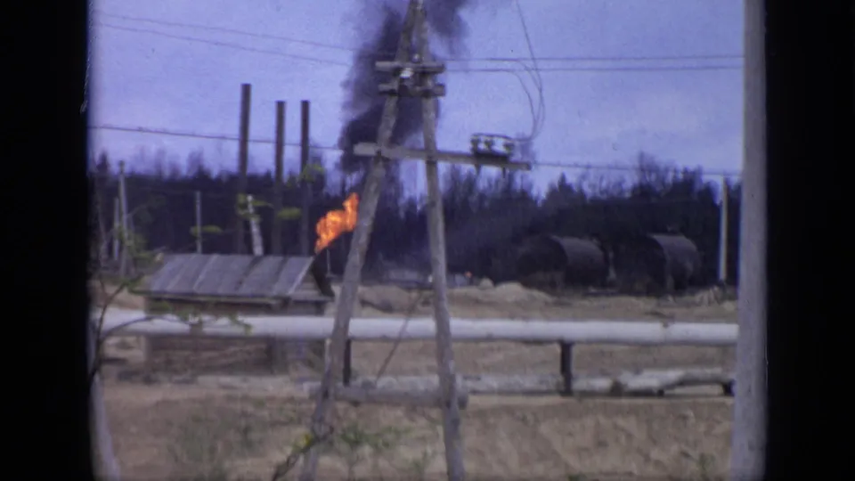 Closer 1971 Moscow-area frame showing orange flames, black smoke, a pole structure, pipeline, dry field, and distant industrial tanks.