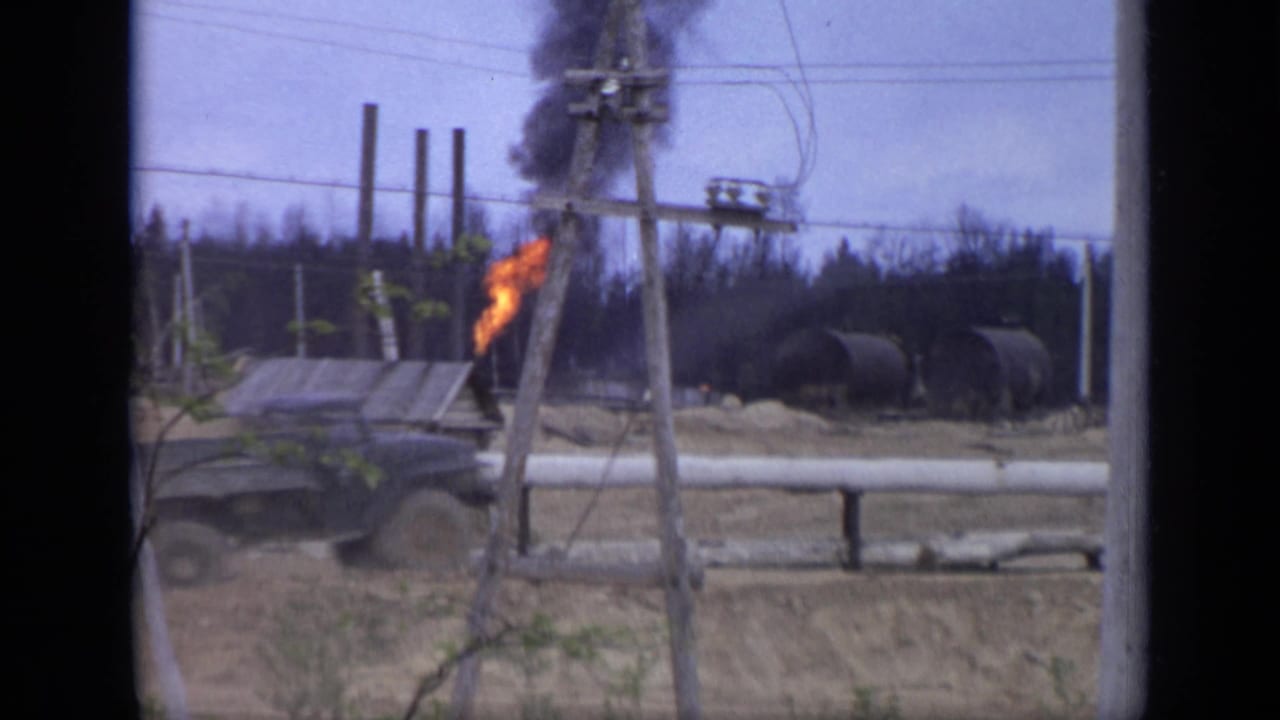 In Moscow, Russia in 1971, a dark truck passes a small grey industrial structure as orange flame and black smoke rise near utility poles.