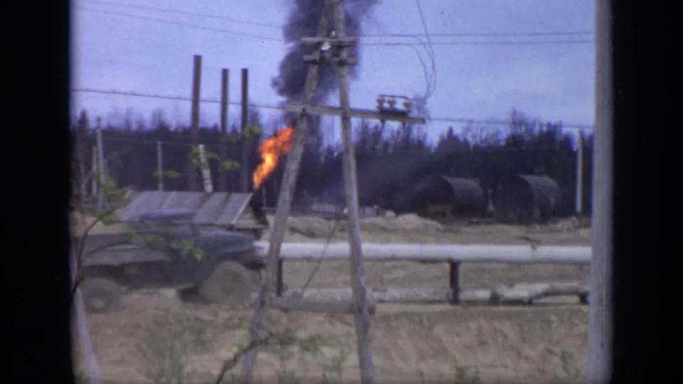 In Moscow, Russia in 1971, a dark truck passes a small grey industrial structure as orange flame and black smoke rise near utility poles.