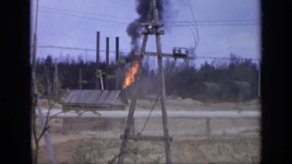 A small grey structure continues to emit orange flame and dark smoke at a Moscow industrial roadside, with pipes and utility wires in view.