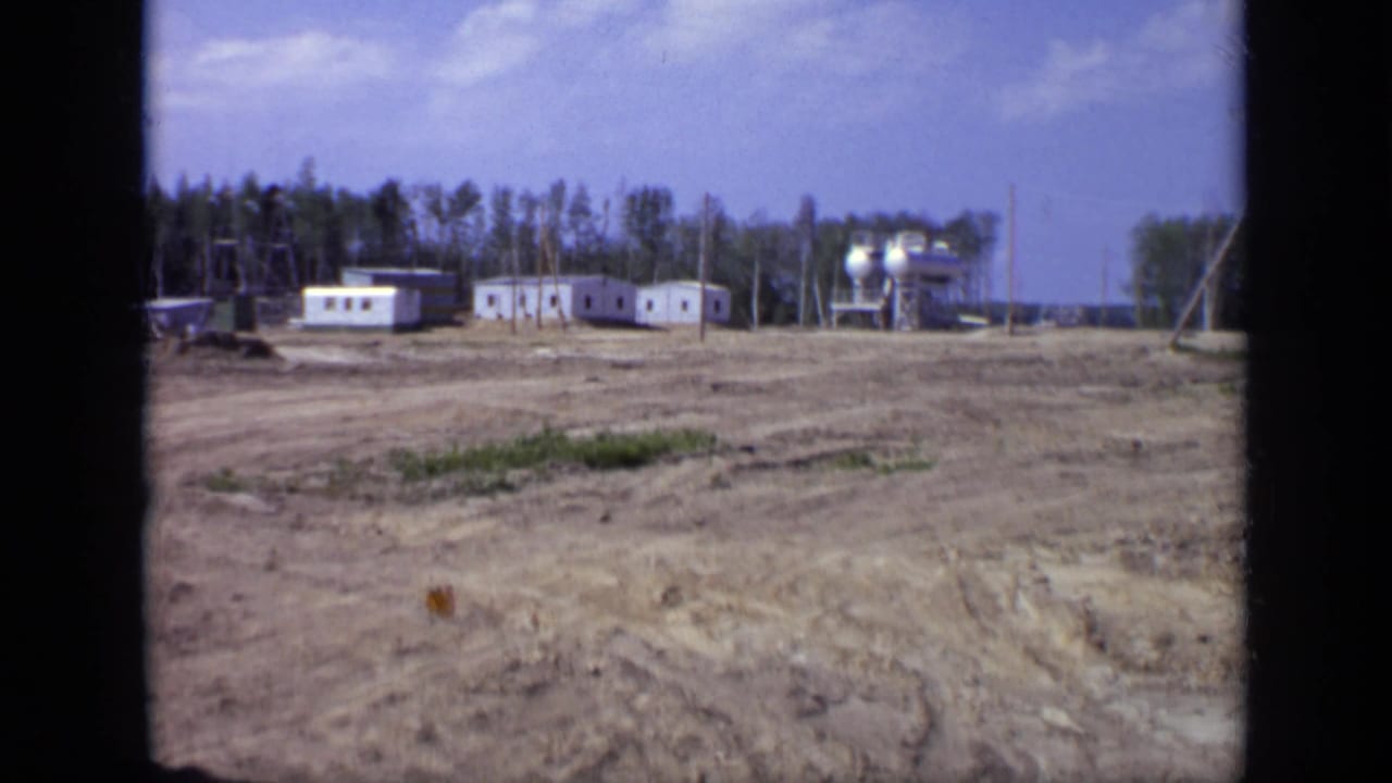 1971 Moscow 8mm frame of a rural industrial worksite with white prefabricated buildings, storage tanks, utility poles, and a wooded backdrop.