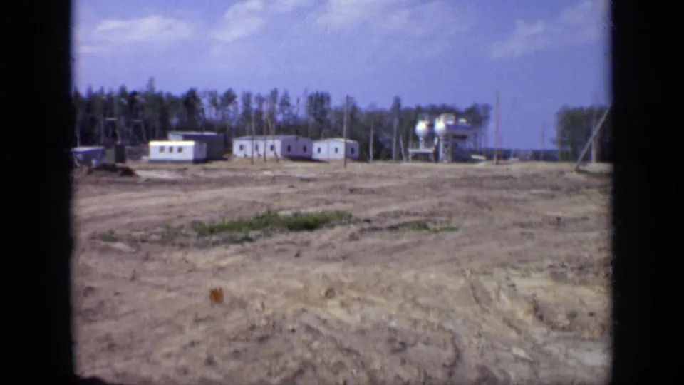 1971 Moscow 8mm frame of a rural industrial worksite with white prefabricated buildings, storage tanks, utility poles, and a wooded backdrop.