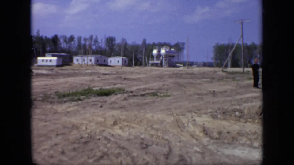 1971 Moscow 8mm frame showing a wide worksite view with prefabricated buildings, tanks, utility poles, trees, and a partly visible person at the edge.
