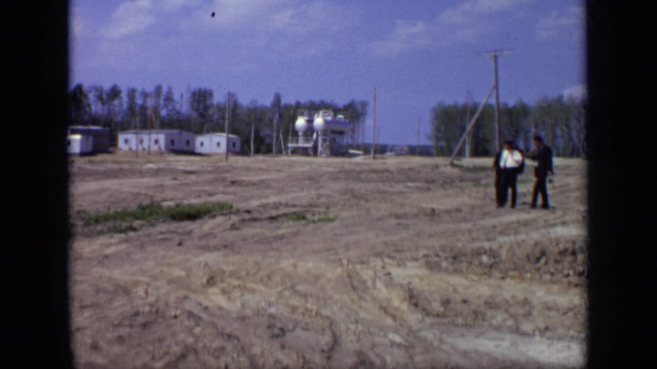 1971 Moscow home-movie frame showing two suited men crossing a bare field near prefab buildings, tanks, and utility poles.