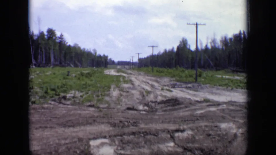 1971 Moscow home-movie view of a rutted dirt track bordered by trees, low vegetation, and a line of utility poles.