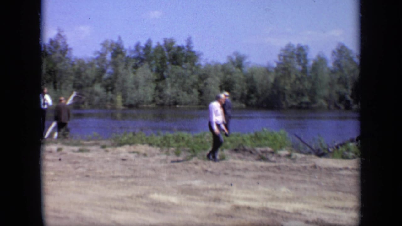 1971 Moscow home-movie frame showing adult men walking along a riverbank with trees across the water.