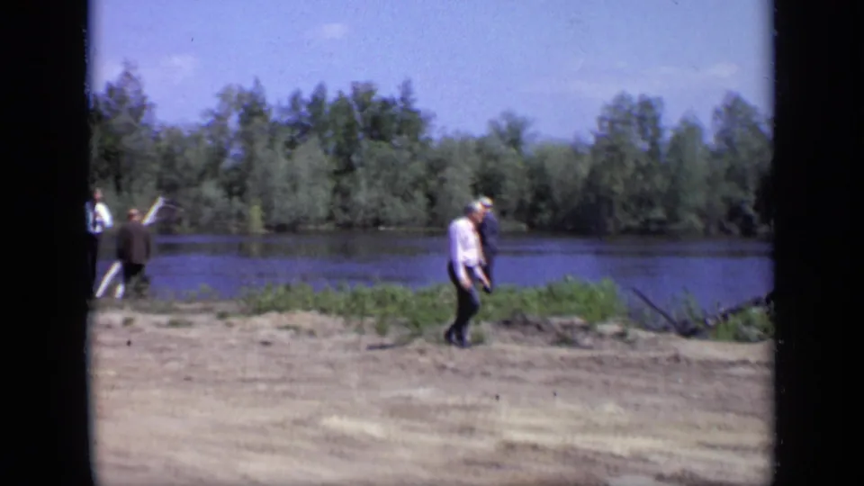 1971 Moscow home-movie frame showing adult men walking along a riverbank with trees across the water.
